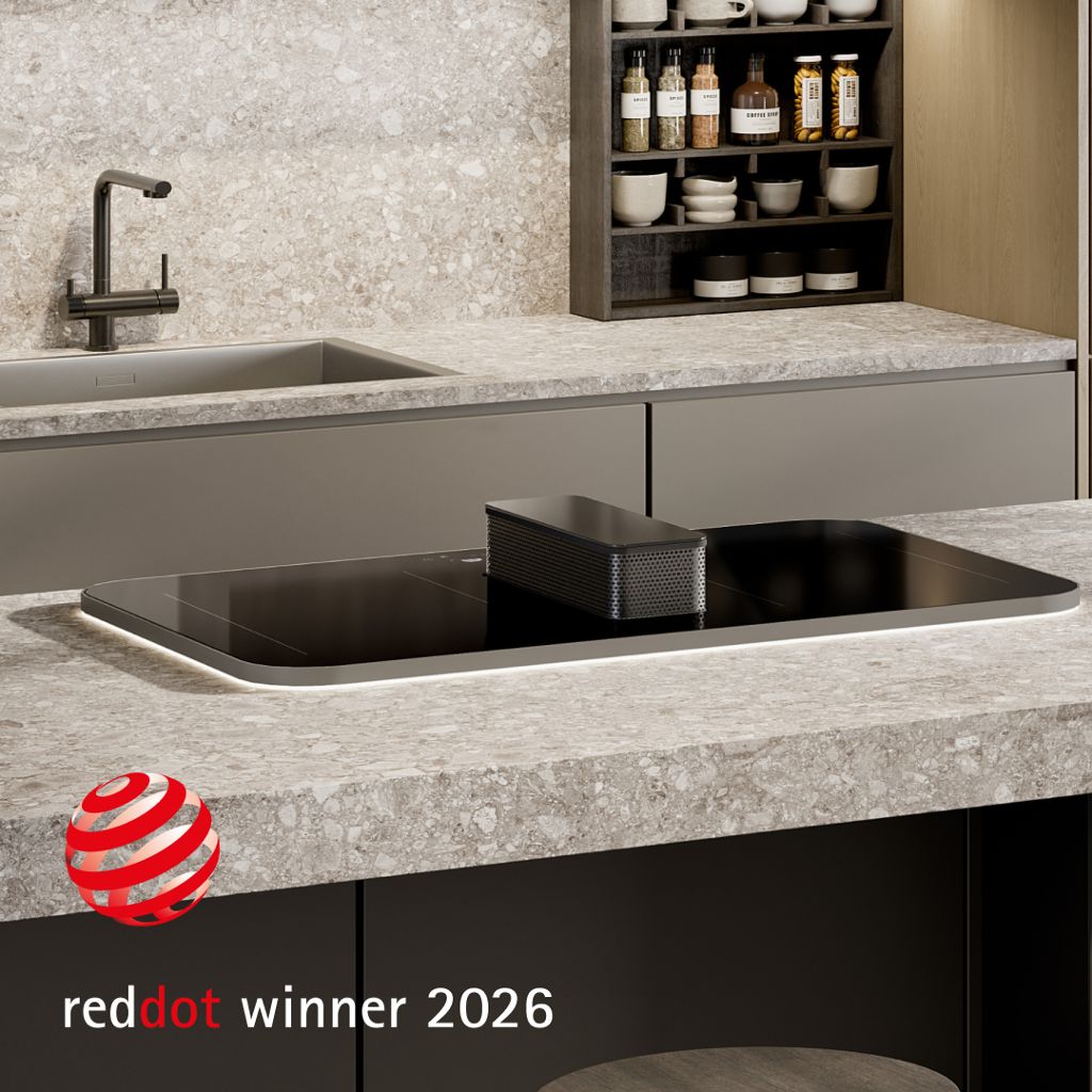 Plaque à induction noire avec hotte aspirante centrale intégrée, installée dans un îlot de cuisine doté d'un plan de travail en pierre claire ; logo « Red Dot Winner 2026 » visible.