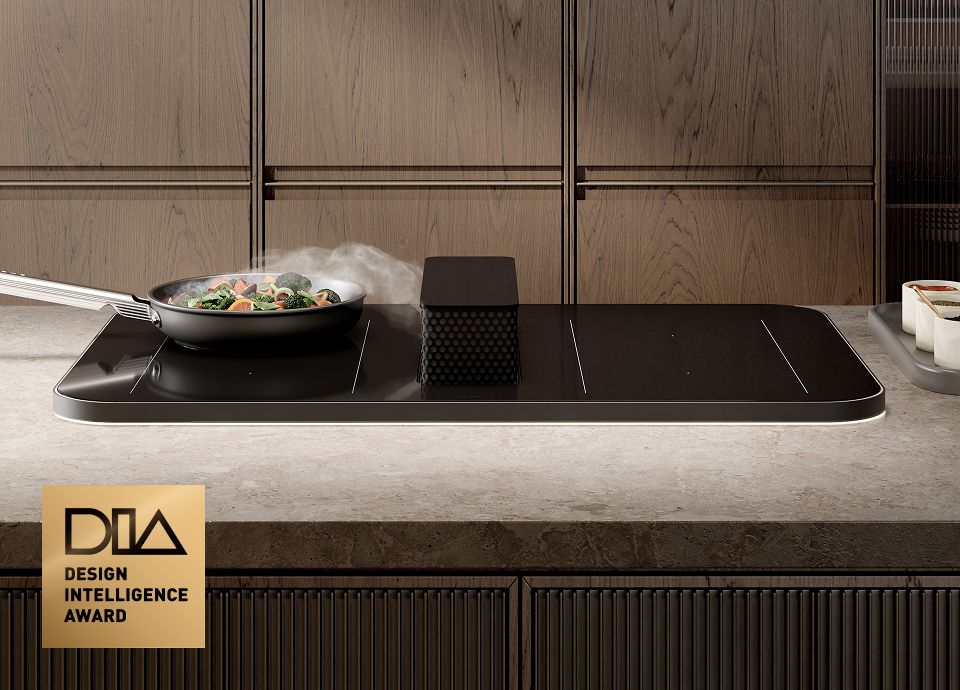 Plaque à induction noire avec hotte intégrée, sur laquelle on voit une casserole dans laquelle cuisent des légumes, de la vapeur s'en échappant, posée sur un plan de travail moderne en pierre, avec des meubles en bois en arrière-plan et un badge du Design Intelligence Award.