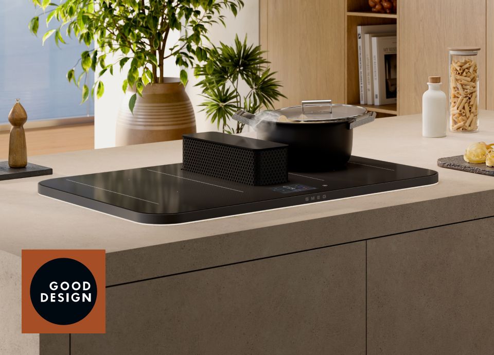 Plaque à induction noire avec hotte centrale intégrée, sur laquelle repose une casserole fermée d'où s'échappe de la vapeur, posée sur un îlot de cuisine moderne agrémenté de plantes, d'armoires en bois et d'accessoires minimalistes, avec un badge du prix Good Design bien visible.