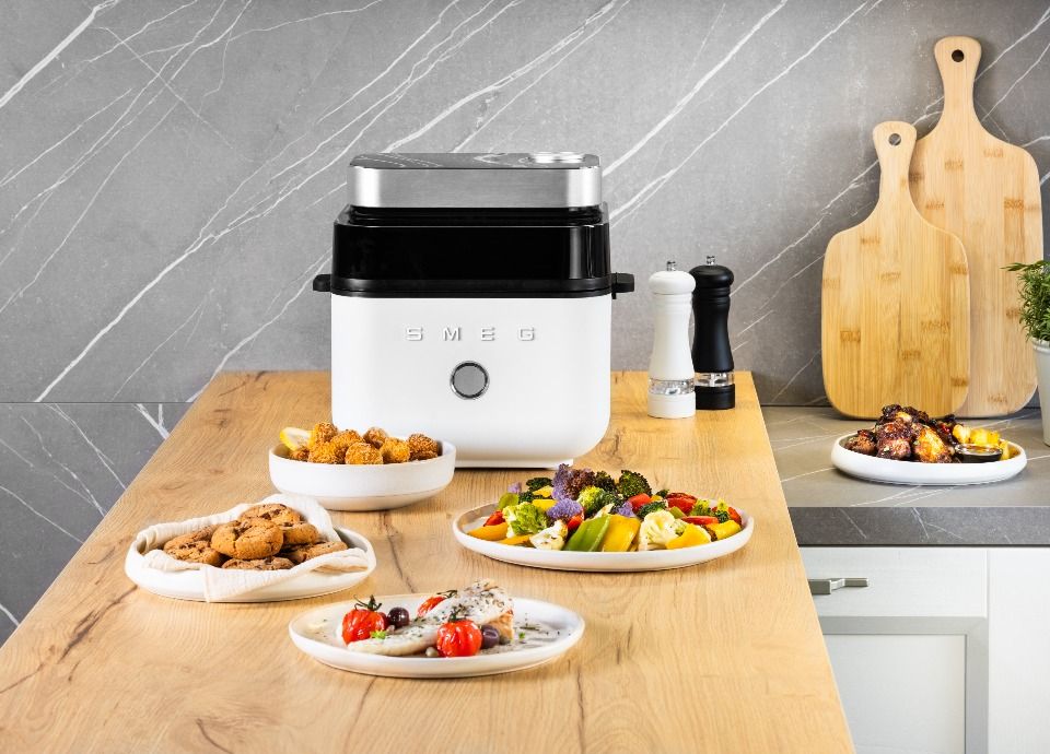 Smeg white air fryer on a wooden countertop surrounded by a variety of dishes including vegetables, chicken and cookies