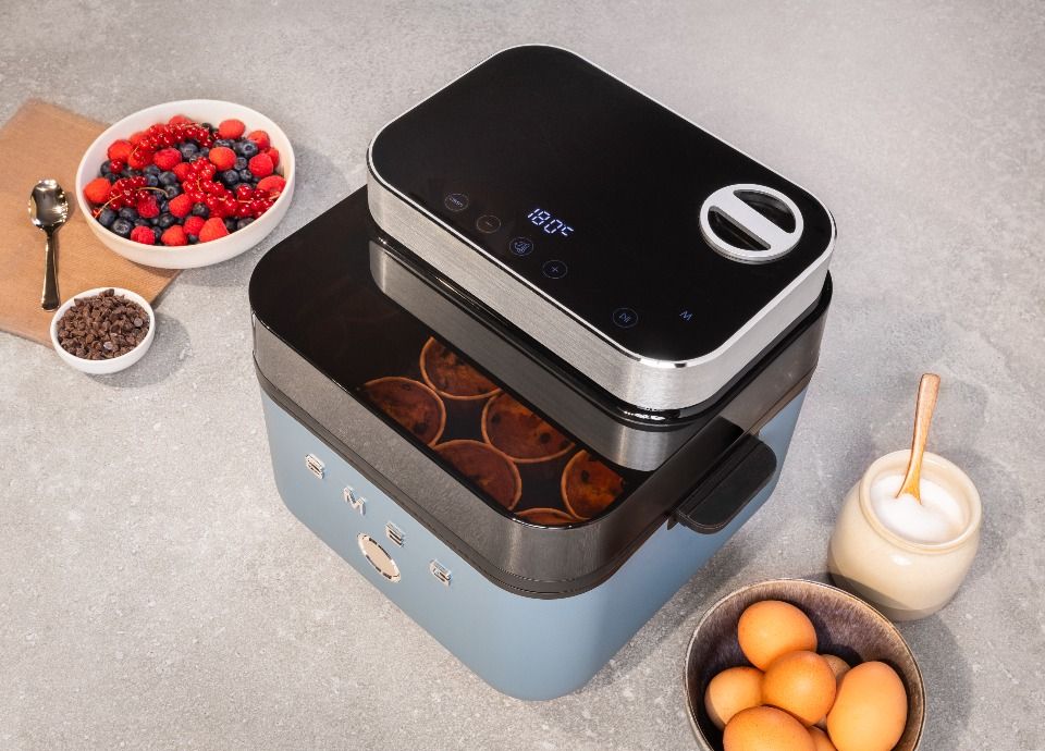 Smeg air fryer in blue finish with transparent window showing muffins, surrounded by eggs, berries and ingredients