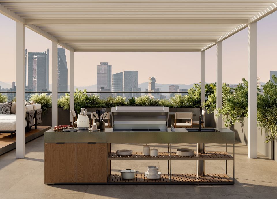 Outdoor kitchen on a terrace covered by a white pergola. In the center, an island with a dark stone countertop and wooden frame with open shelves holding plates and bowls. On the counter, kitchen utensils and a built-in stainless steel barbecue with the lid open. On the right, a sink with a chrome tap. In the background, green plants along the edge and a panoramic view of city skyscrapers.