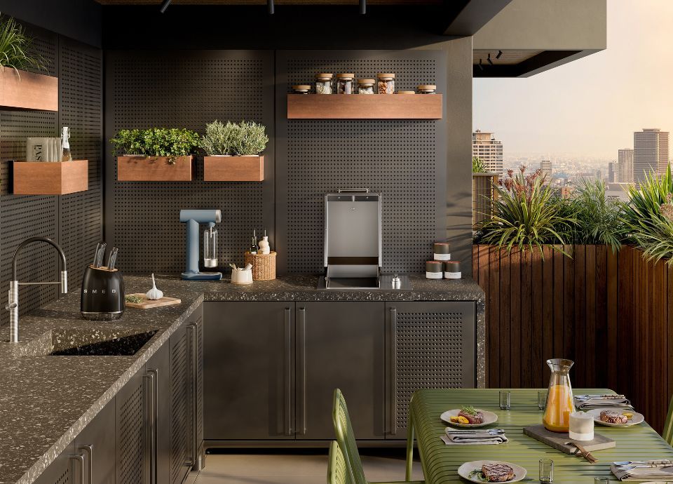 Modern outdoor kitchen with a dark stone countertop and cabinets featuring perforated panel doors. In the center of the counter, there is a built-in stainless steel barbecue with the lid open in a vertical position. On the left side, there is a sink with a chrome faucet and a storm blue soda maker. Wooden shelves on the back wall hold jars of spices and potted herbs. In the foreground, a table is set with a green tablecloth, plates, glasses, and a jug of juice. In the background, a balcony with plants offers a panoramic view of the city.