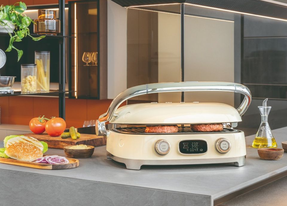 Een SMEG Multifunctionele Grill in crème afwerking die burgers grillt op een eigentijds keukeneiland.