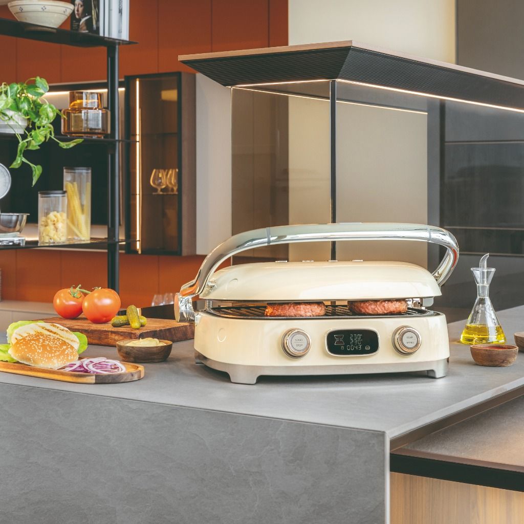 Tostadeira Multiuso Smeg em creme a preparar hambúrgueres sobre uma ilha de cozinha contemporânea