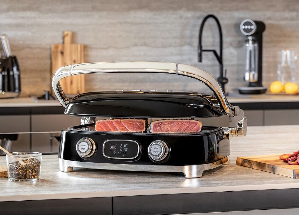 Black multi-use grill with an open lid cooking steaks on a kitchen countertop, featuring dual control knobs and a digital display in a modern kitchen setting.