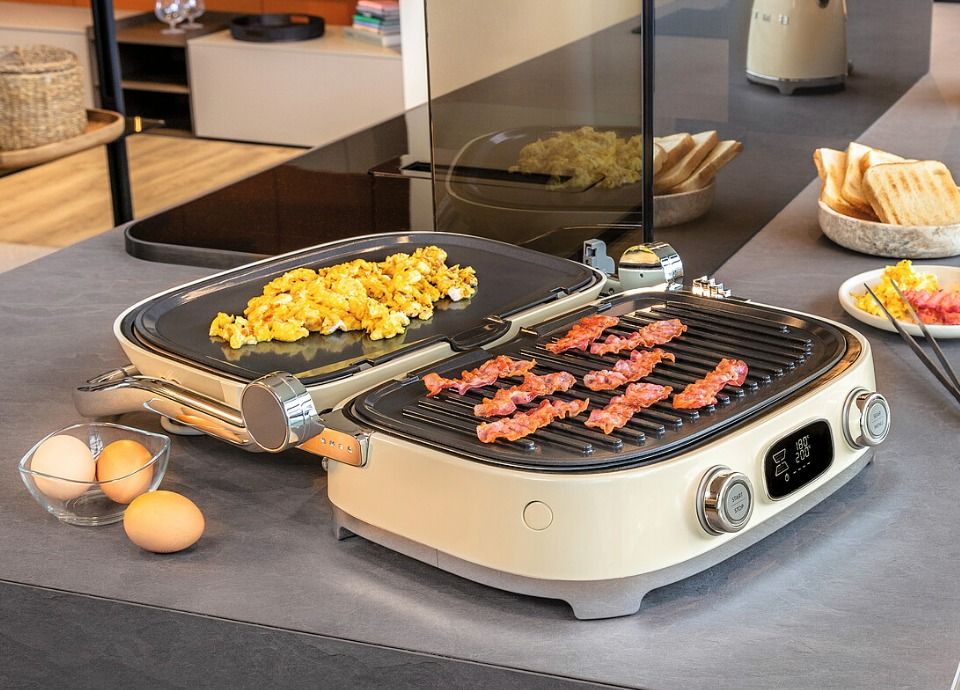 Cream multi-use grill with open lid cooking eggs and bacon on interchangeable plates, placed on a modern kitchen island with bread, coffee cups and breakfast setup in the background.