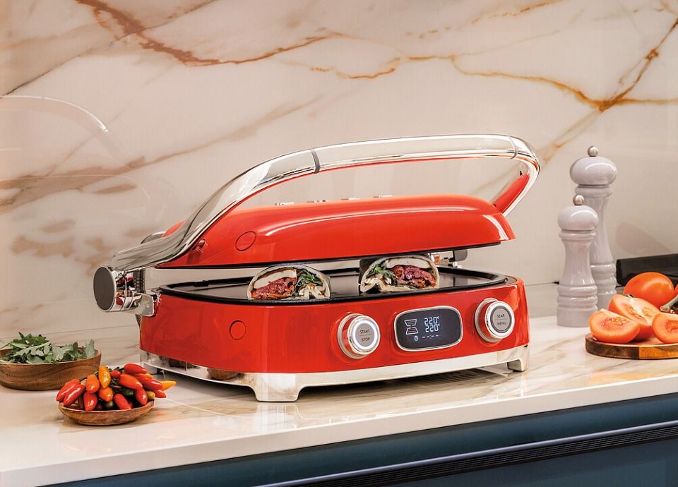 Red multi-use grill with chrome handle and digital controls on a kitchen countertop, surrounded by fresh vegetables, a cutting board and a marble backsplash in a modern kitchen.
