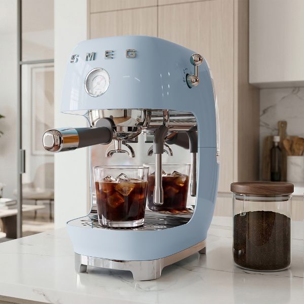 Cafetera Espresso Manual Smeg en color azul pastel en una cocina en tonalidades crema y blanco. En la cafetera hay un vaso con hielo y café, mientras la cafetera está dispensando café en función Cold Brew.