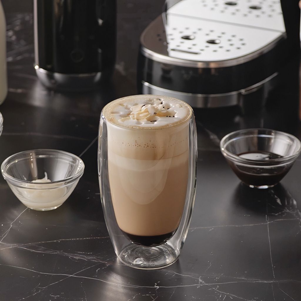 A layered choco coconut latte macchiato served in a double‑walled glass, topped with whipped foam and coconut flakes.