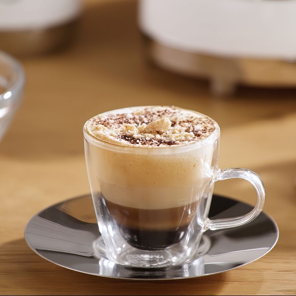 A layered choco macchiato served in a clear glass cup on a reflective saucer, topped with creamy foam and chocolate crumbles.