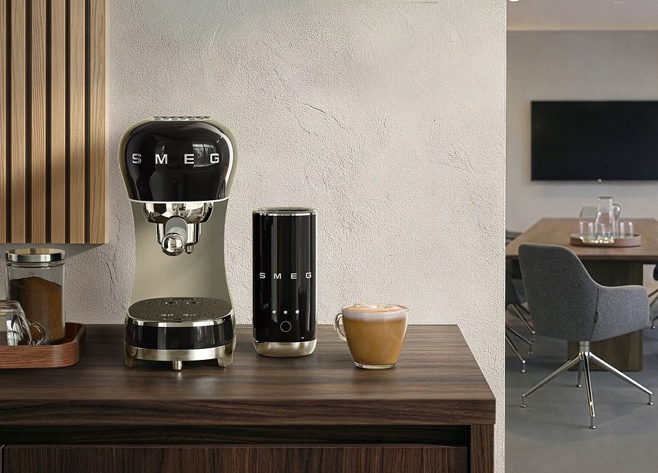 Black SMEG espresso coffee machine and matching milk frother shown together as a coffee maker set on a wooden countertop with a cappuccino cup and coffee grounds jar in a modern kitchen setting
