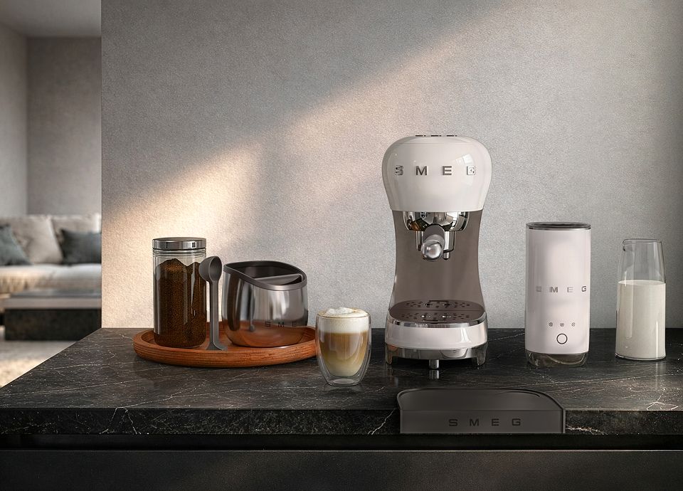 White SMEG espresso machine with matching milk frother coffee set on a dark marble countertop with a latte glass, milk carafe, coffee grounds jar, and coffee accessories in a modern kitchen setting