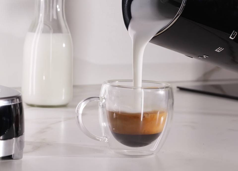 ream SMEG espresso machine with matching milk frother coffee set on a wooden countertop with a latte glass, milk carafe, and coffee grounds jar in a modern kitchen setting