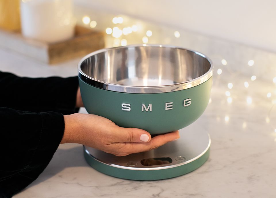 Hands holding an emerald green SMEG kitchen scale, resting on a marble countertop. Blurred decorative lights in the background.