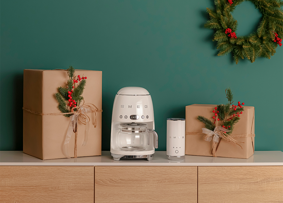 Filter coffee machine and milk frother by SMEG, both in cream color, placed on a light wooden cabinet, flanked by two gift boxes wrapped in brown paper and decorated with pine branches and red berries. In the background, a green wall with a Christmas wreath.
