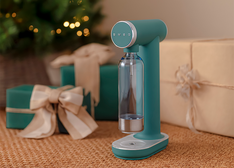 Emerald green SMEG water carbonator with a transparent bottle, placed on a rug in front of gift boxes wrapped in beige and green paper and decorated with bows. In the background, a lit Christmas tree.