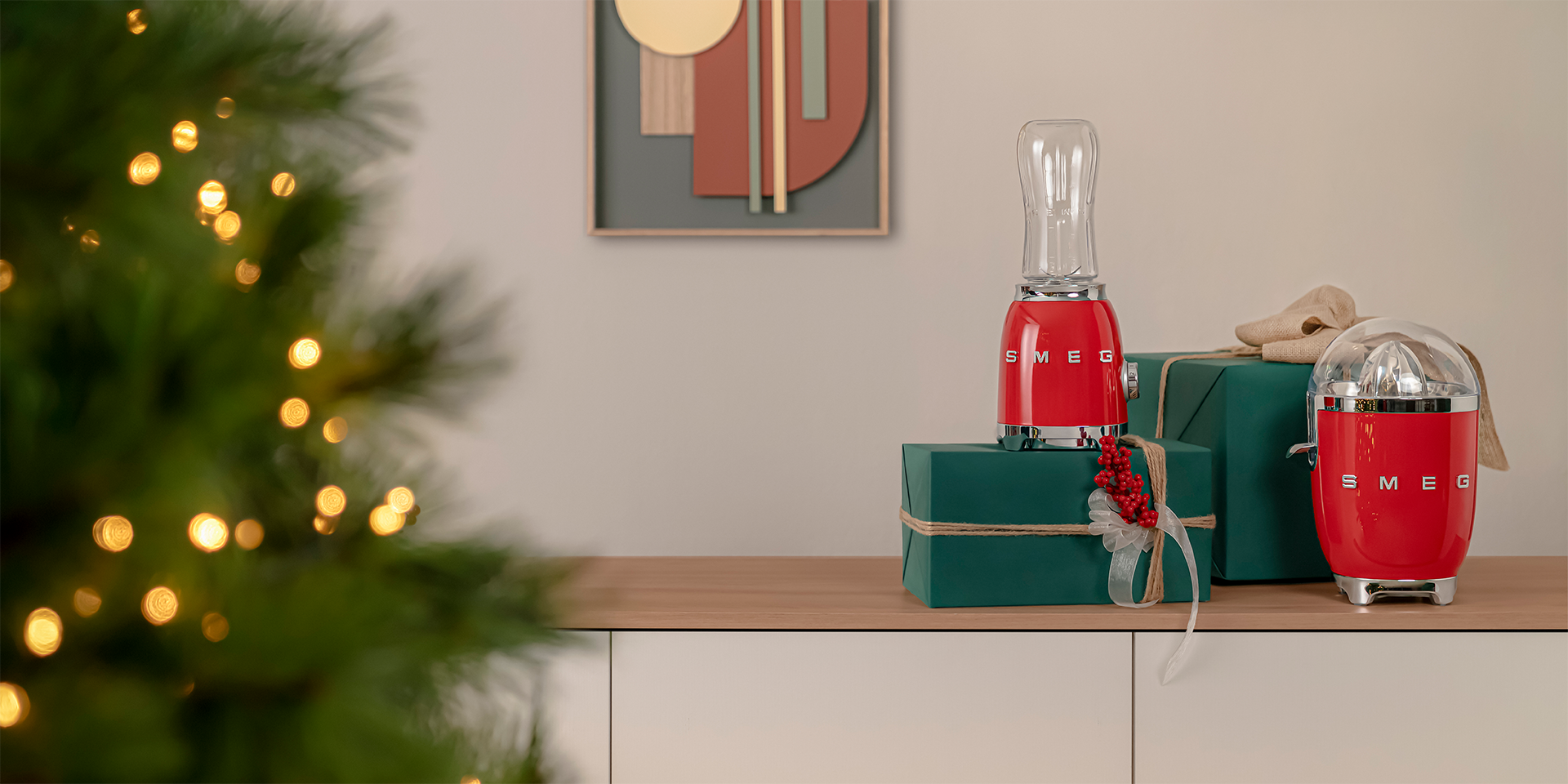 Red Personal Blender and Citrus Juicer with wrapped boxes in green wrapping paper. The background is neutral and in the left corner is part of a Christmas tree with fairy lights