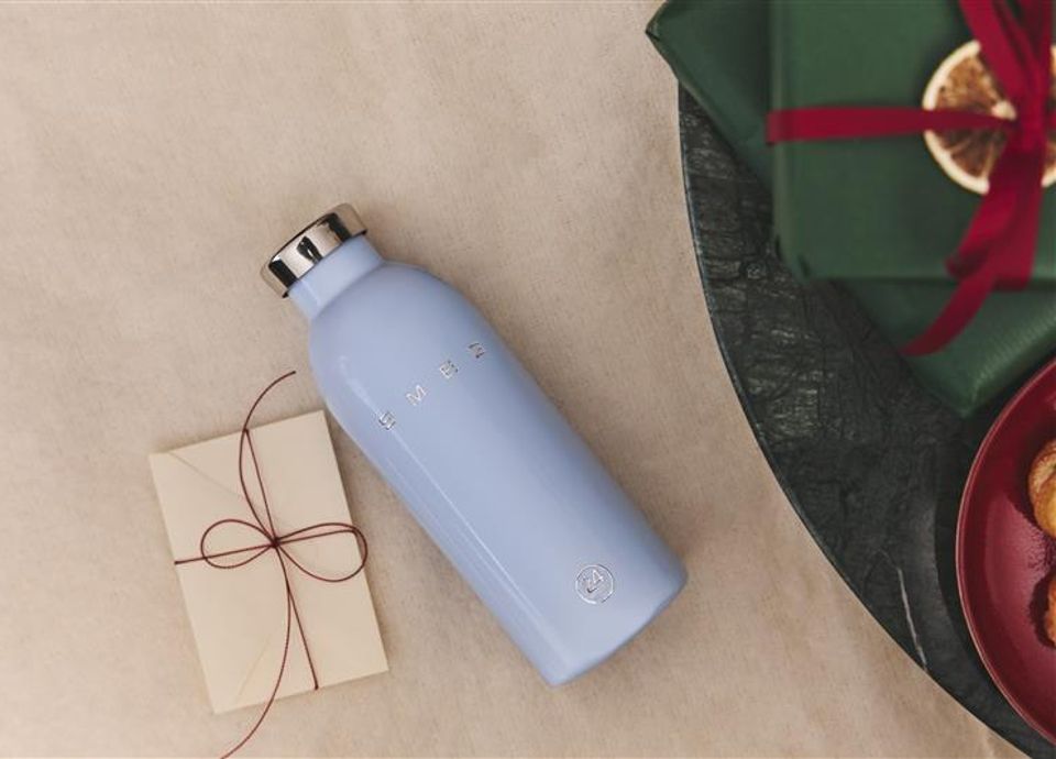 Light blue stainless steel bottle with chrome cap and SMEG logo, placed on a light surface next to an envelope tied with red string. Partially visible green gift boxes with red ribbons.