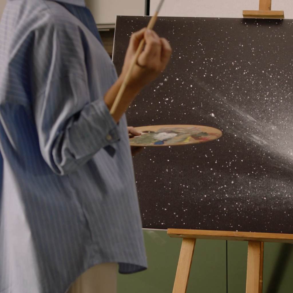 Artist holding a paintbrush and palette while painting a galaxy-inspired canvas on an easel