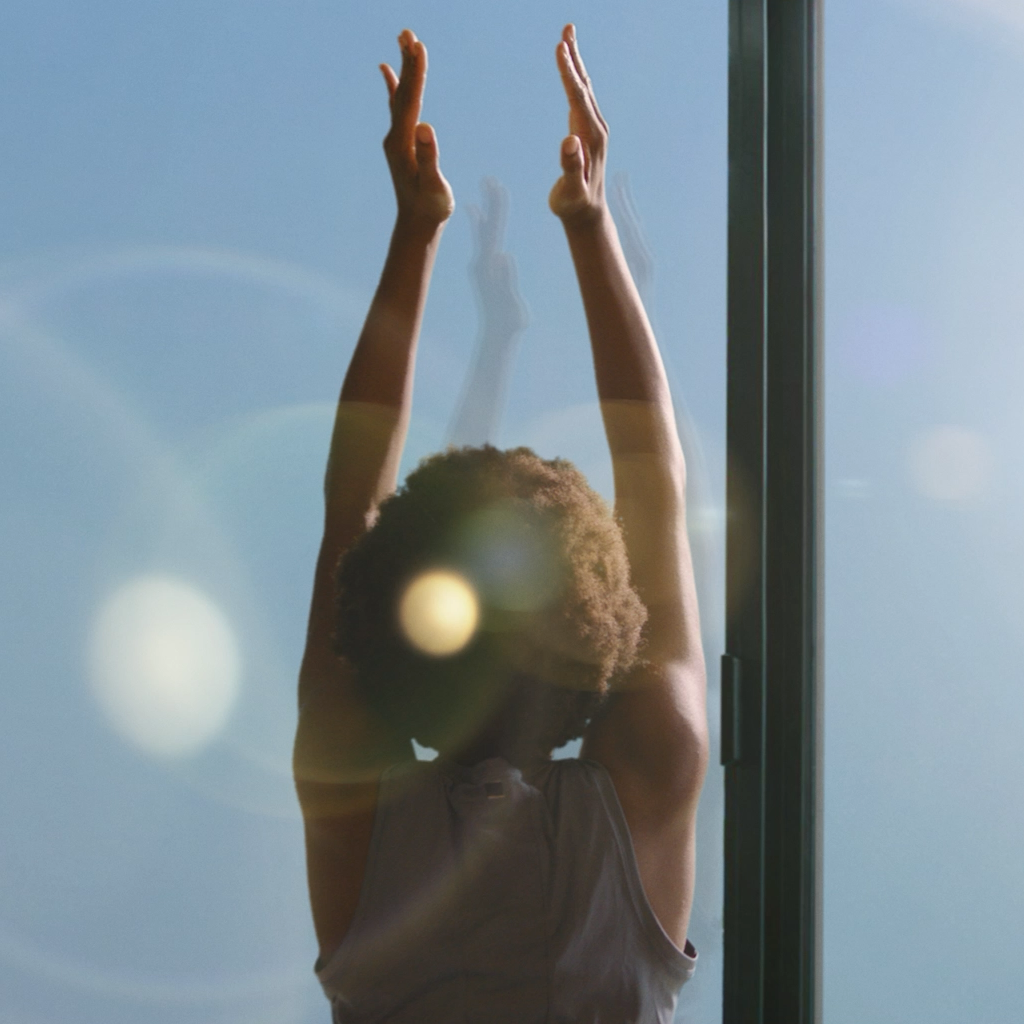 Backlit figure stretching arms upward near a window with sunlight and lens flare effects