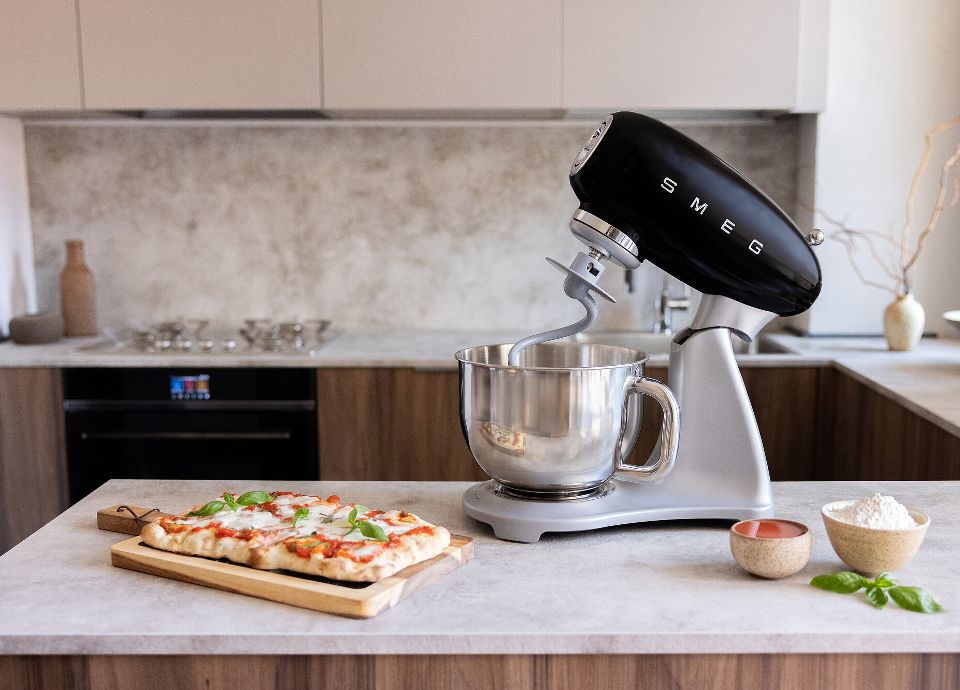 Robot sur socle SMEG noir avec corps en métal et bol en acier inoxydable posé sur le plan de travail de la cuisine. À côté, sur le plan de travail, se trouve un plateau à pizza garni de tomates, de mozzarella et de feuilles de basilic frais. Il y a également trois petits bols : un avec de la farine blanche, un avec des tomates et un avec des feuilles de basilic. À l'arrière-plan, on peut voir une cuisine en bois clair avec des armoires murales beiges, un four SMEG encastré, une plaque de cuisson SMEG en acier inoxydable et quelques objets décoratifs.