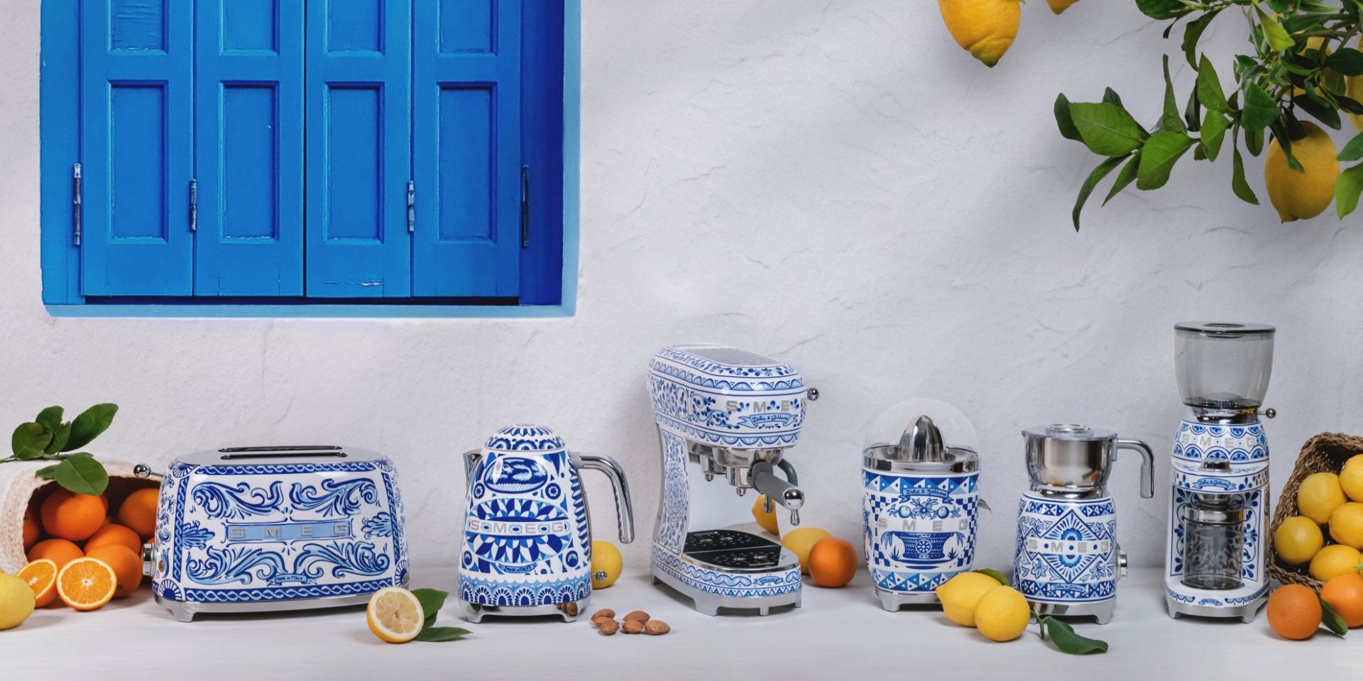 Un grille-pain, une bouilloire, une machine à café expresso, un presse-agrume, une chocolatière et un broyeur à café de la gamme Blu Mediterraneo en collaboration avec Dolce&Gabbana sont allignés sur une table blanche.