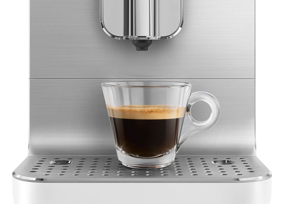 Glass cup of espresso placed on the drip tray of a stainless steel Smeg coffee machine in a minimalist setting