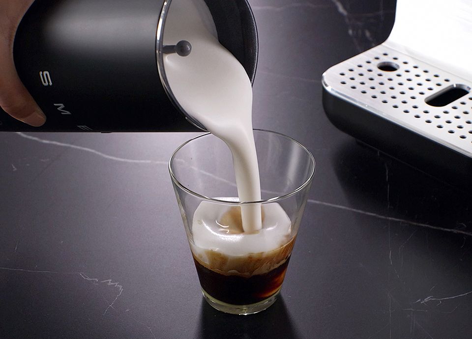 Frothy milk being poured from a SMEG milk frother into a glass of coffee on a dark countertop.