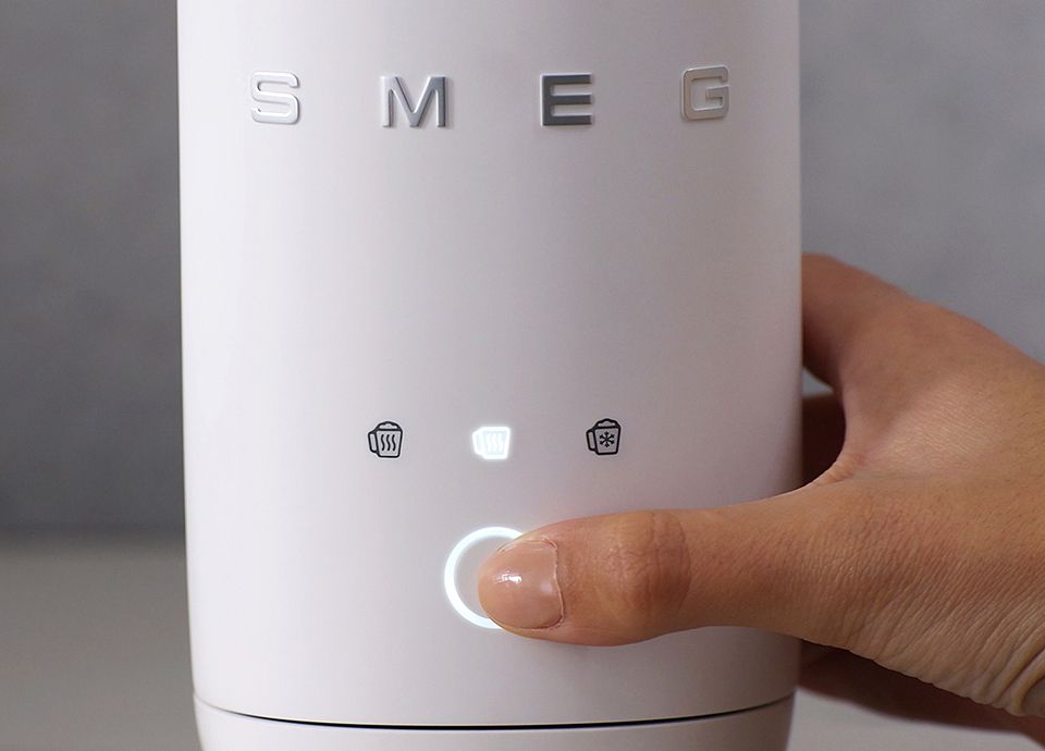 Close-up of a SMEG milk frother with illuminated touch control button being pressed and function icons displayed above.