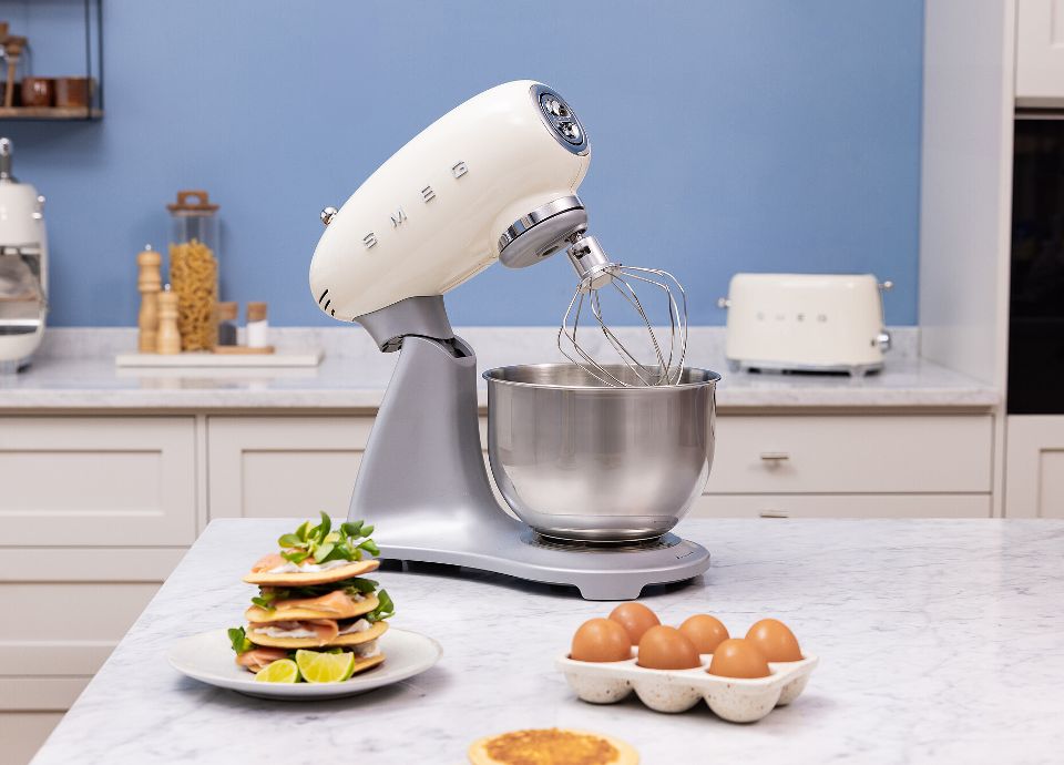 On a marble kitchen countertop, there is a white SMEG Stand Mixer with stainless steel body. Next to the SMEG Stand Mixer, there are some eggs in an egg cup. In the foreground, on a plate, there is a small stack of pancakes filled with vegetables and lemon slices. In the blue background, you can see other white SMEG kitchen appliances and spice jars. The room is tidy and bright.