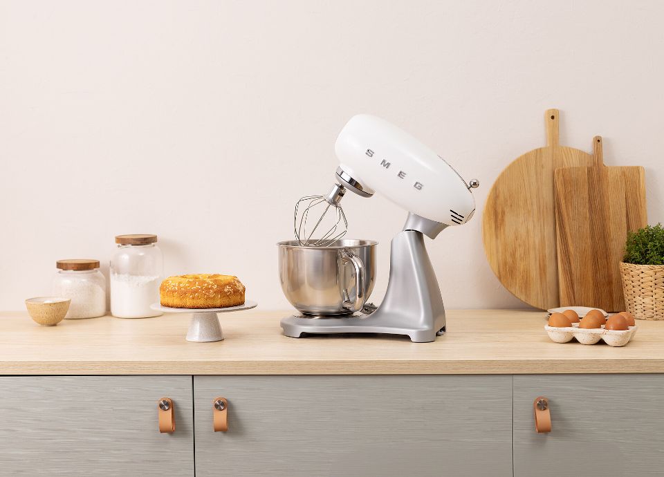 On a light-colored kitchen countertop, there is a white SMEG Stand Mixer with stainless steel body, a baked dessert on a cake stand, two glass jars containing sugar and flour, some eggs in a container, and a few wooden cutting boards leaning against the wall. The room is tidy and bright.