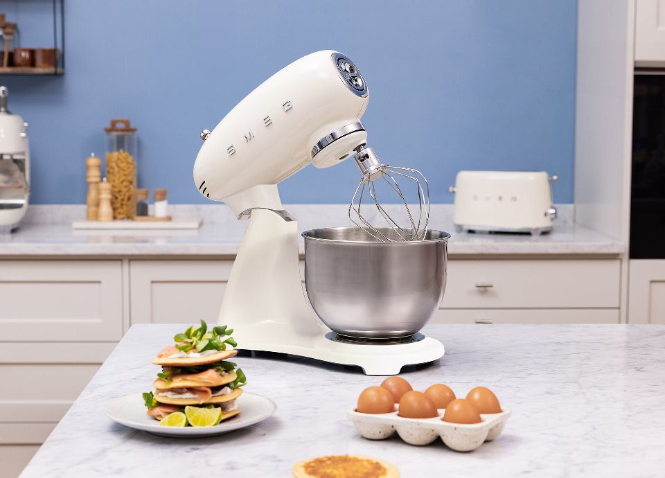 On a marble kitchen countertop, there is a white SMEG Stand Mixer with stainless steel body. Next to the SMEG Stand Mixer, there are some eggs in an egg cup. In the foreground, on a plate, there is a small stack of pancakes filled with vegetables and lemon slices. In the blue background, you can see other white SMEG kitchen appliances and spice jars. The room is tidy and bright.