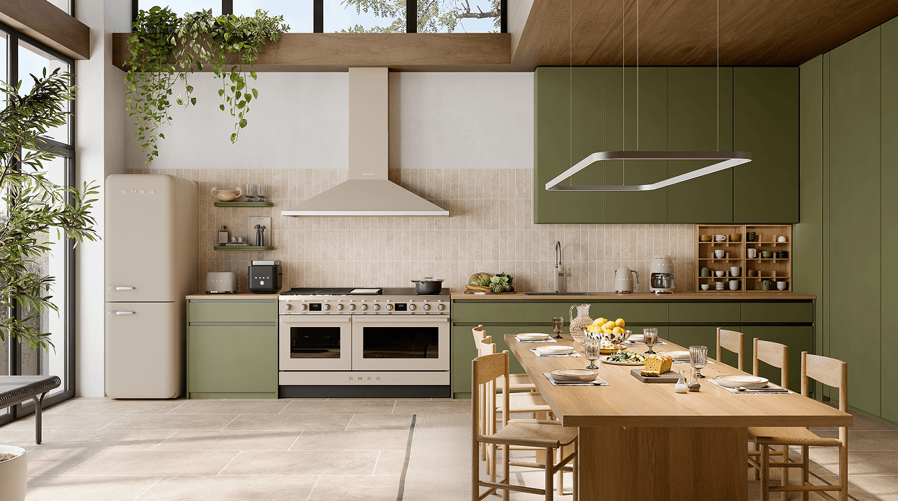 Bright contemporary kitchen featuring SMEG appliances in matt moonlight finish, including a range cooker, extractor hood, and retro-style refrigerator, set within green cabinetry, warm wood accents, and a large dining table under natural light.