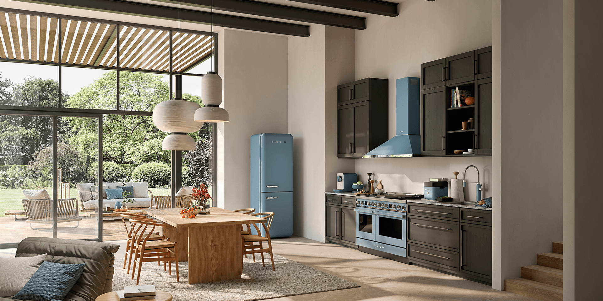 Spacious modern kitchen with SMEG appliances in matte storm blue finish, including a retro-style refrigerator, range cooker, and extractor hood, set within dark cabinetry, a light wooden dining table, and large windows opening onto a garden.
