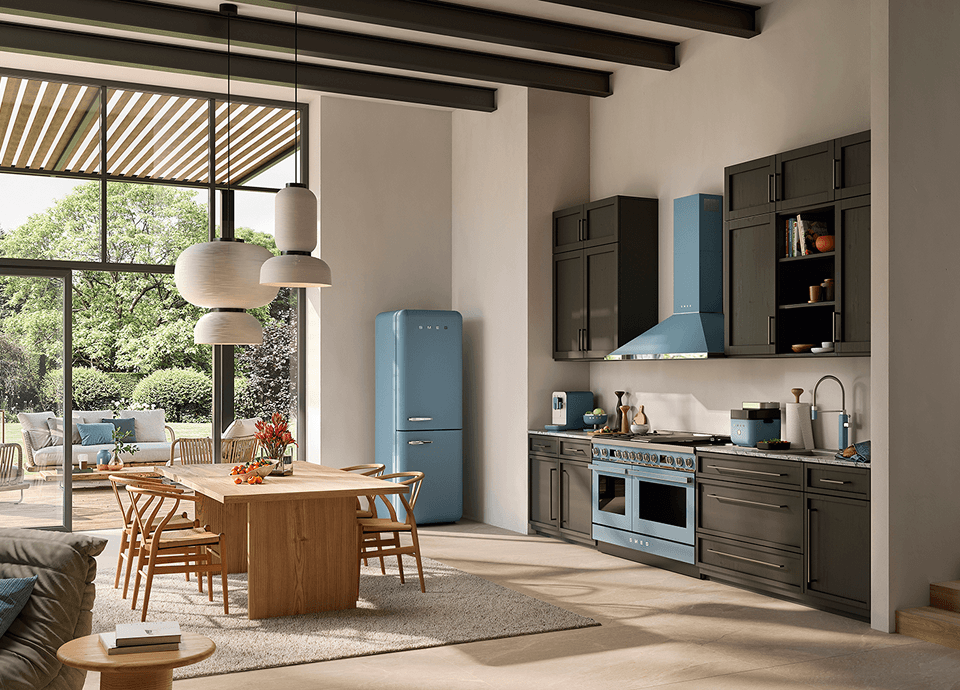 Cuisine spacieuse et moderne équipée d'appareils électroménagers SMEG au fini bleu tempête mat, comprenant un réfrigérateur, un piano de cuissonet une hotte aspirante de style rétro, le tout intégré dans des meubles de cuisine foncés, avec une table à manger en bois clair et de grandes fenêtres donnant sur le jardin.