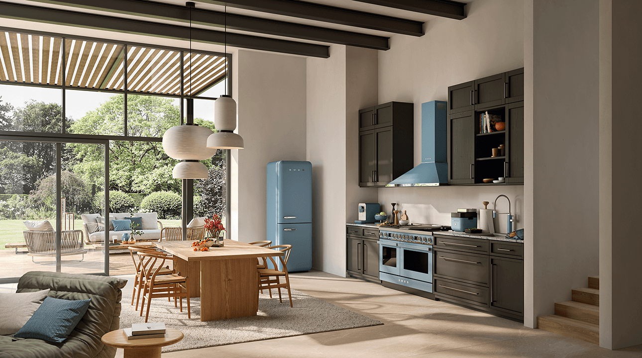 Spacious modern kitchen with SMEG appliances in matte storm blue finish, including a retro-style refrigerator, range cooker, and extractor hood, set within dark cabinetry, a light wooden dining table, and large windows opening onto a garden.