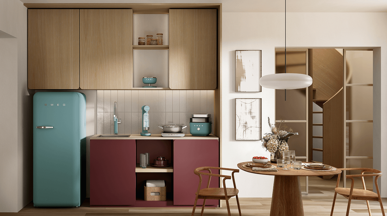 Compact kitchen corner featuring SMEG appliances in storm blue finish, including a retro-style refrigerator, soda maker, and small countertop appliances, set against warm wood cabinetry and burgundy lower units with a dining area to the side.