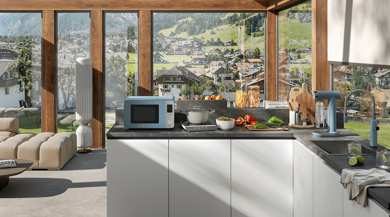 Contemporary kitchen with large floor-to-ceiling windows overlooking a green landscape, featuring SMEG appliances and accessories with storm blue accents on a central island, surrounded by dark cabinetry, wood ceilings, and natural light.
