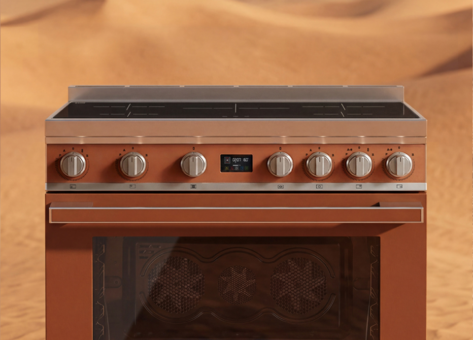 Freestanding SMEG cooker in matt amber clay colour, featuring stainless steel control knobs and a black glass oven door, set against a desert landscape with soft sand dunes in the background.