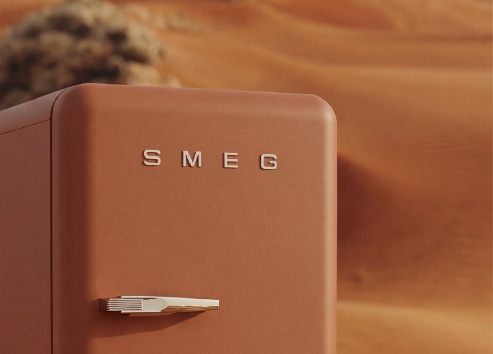 SMEG Fab refrigerator in matt amber clay colour, featuring stainless steel control knobs and a black glass oven door, set against a desert landscape with soft sand dunes in the background.