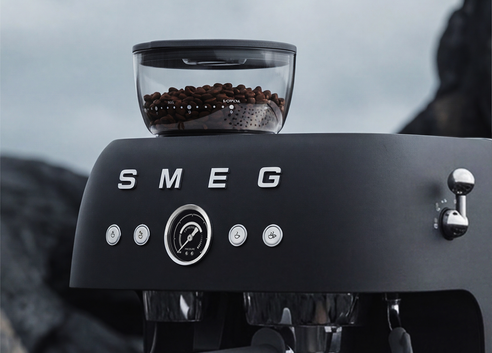 Close-up of a SMEG espresso coffee machine in matt black, featuring a built-in grinder with visible coffee beans, a central pressure gauge, and chrome control details, set against a dark rocky outdoor background.