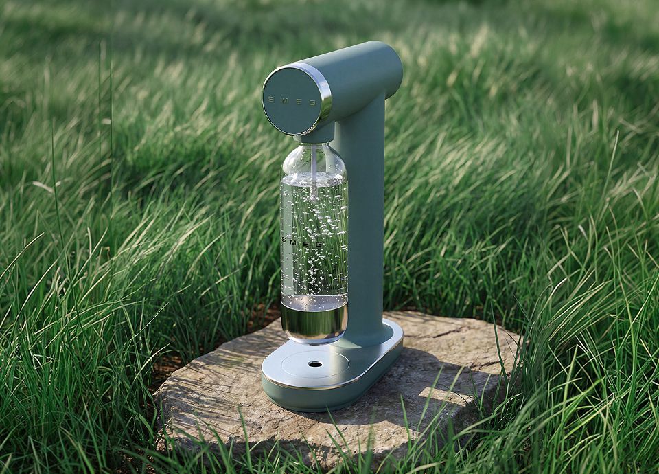 Emerald green SMEG soda maker with reusable sparkling water bottle, displayed on a natural stone surface surrounded by lush green grass in an outdoor setting.