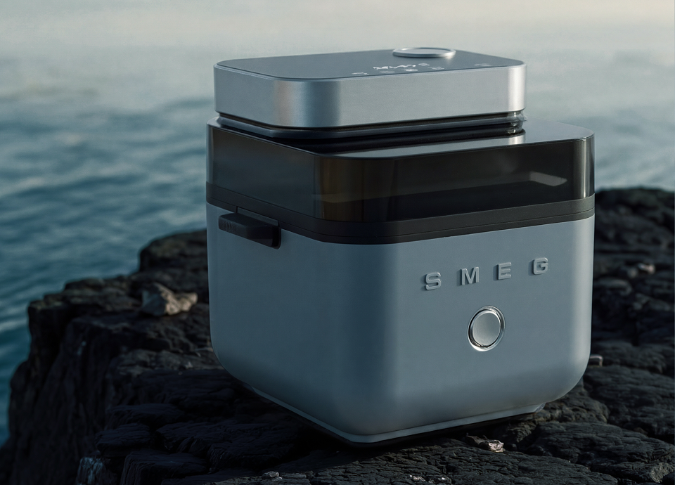SMEG air fryer in storm blue colour, featuring a compact square design with a dark glass front panel and metallic top, placed on a rocky coastal surface with the sea in the background.