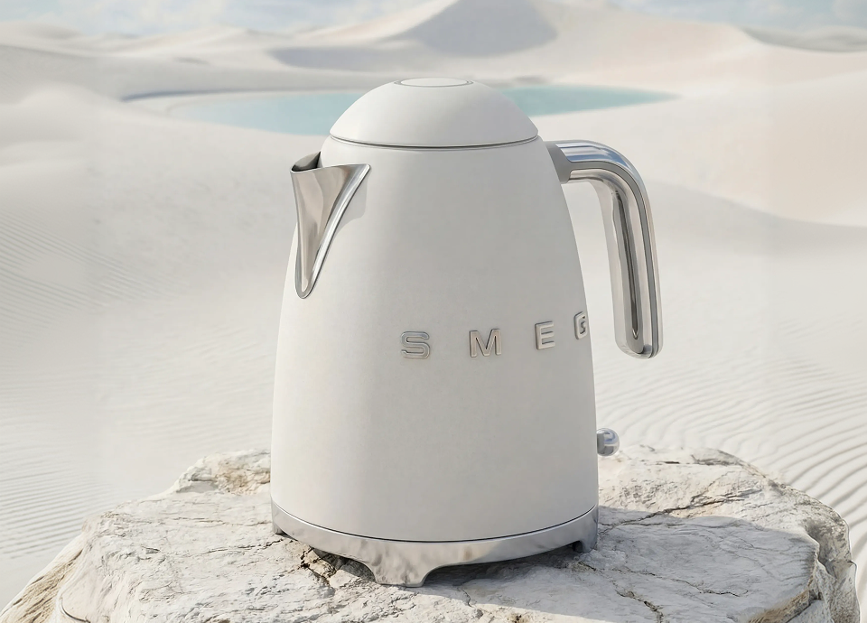 SMEG electric kettle in white matt finish, featuring a smooth curved body with chrome handle and spout, placed on a textured rock surface within soft sand dunes and a calm water backdrop.