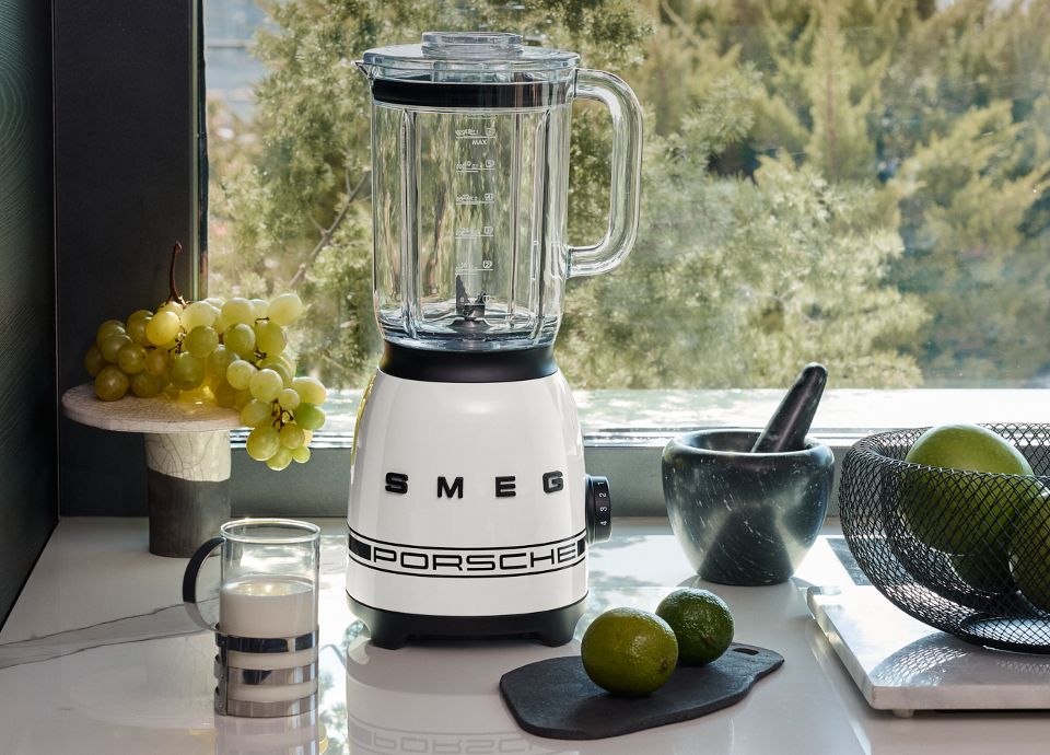Un interior de sala de estar moderno y elegante. En el centro se encuentra un frigorífico SMEG blanco, flanqueado por una planta en maceta y un cuadro apoyado contra la pared. Frente al frigorífico hay dos sillones beige, cada uno con cojines de colores; uno de ellos tiene una manta verde colocada encima. Entre los sillones hay una mesa baja con varios libros. La habitación está bañada por luz natural que entra a través de grandes ventanales cubiertos con cortinas translúcidas, que ofrecen la vista de un coche deportivo aparcado en el exterior. La disposición es limpia y accesible, con un contraste visual claro y sin elementos obstructivos.