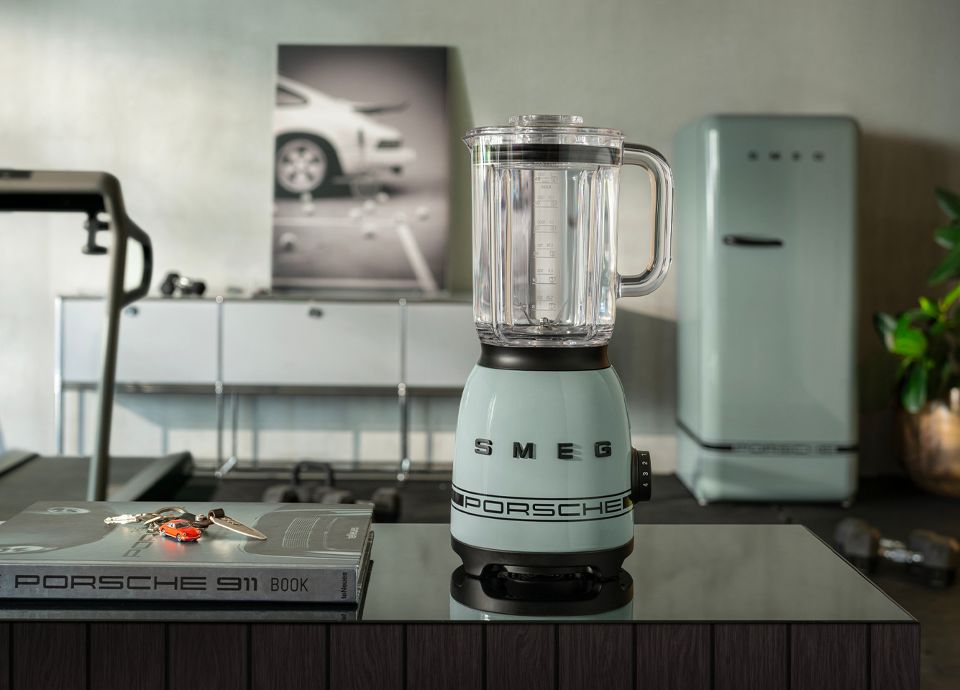 Un espacio moderno con paneles de madera en las paredes y una mesa larga rodeada de sillas. En un lado de la habitación hay un frigorífico Porsche x SMEG Shade Green y un aparador que muestra un hervidor Porsche x SMEG Shade Green, libros, una lámpara y pequeños objetos decorativos. Grandes ventanales ofrecen una vista panorámica de la vegetación, un coche Porsche 911 GT3 RS aparcado en el exterior y el océano al fondo. La disposición es amplia y bien iluminada con luz natural, garantizando una visibilidad clara y accesibilidad para usuarios con discapacidad visual.
