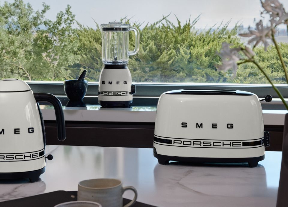 Encimera de cocina con tres electrodomésticos de la colección Porsche x SMEG Carrara White. Los tres electrodomésticos están dispuestos de forma frontal y con un fondo de vegetación verde.