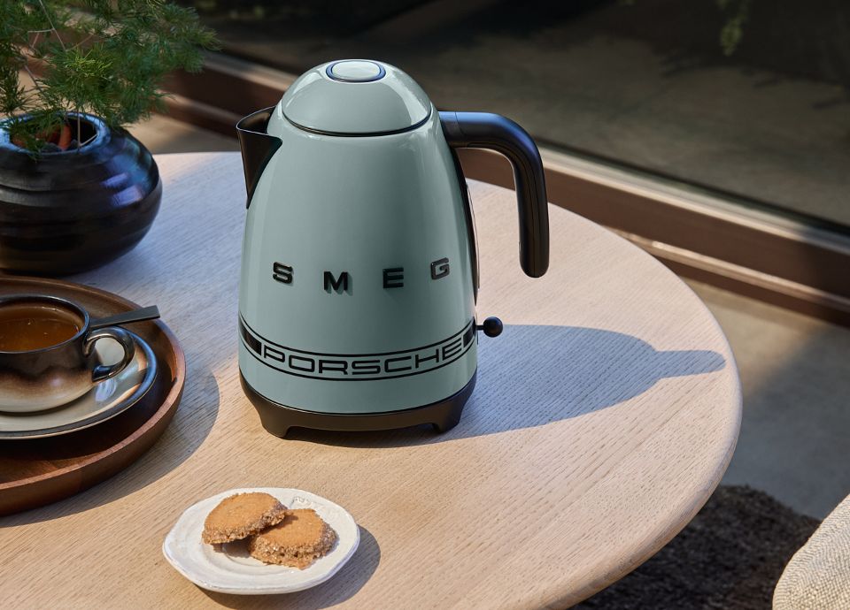 KLF03PSGAU Shade Green Porsche x SMEG kettle on a coffee table with a glass of tea and a cookie