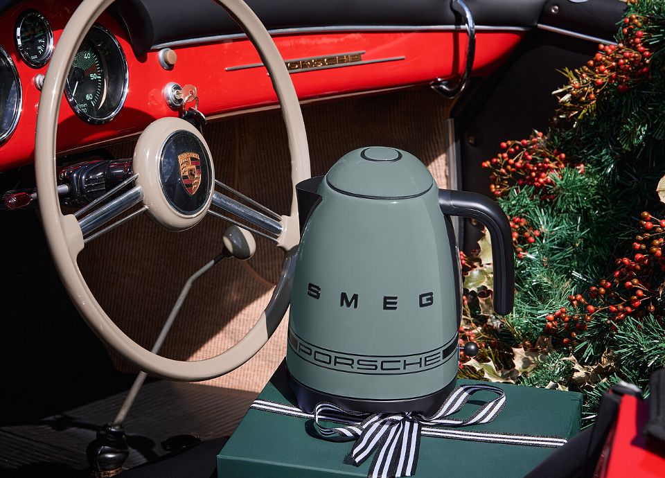 Interior of a vintage Porsche sports car with a light steering wheel and red dashboard. On the seat, there is a SMEG dark green kettle with logo Porsche, placed on a green gift box tied with a black-and-white ribbon. In the background, Christmas decorations with red berries.
